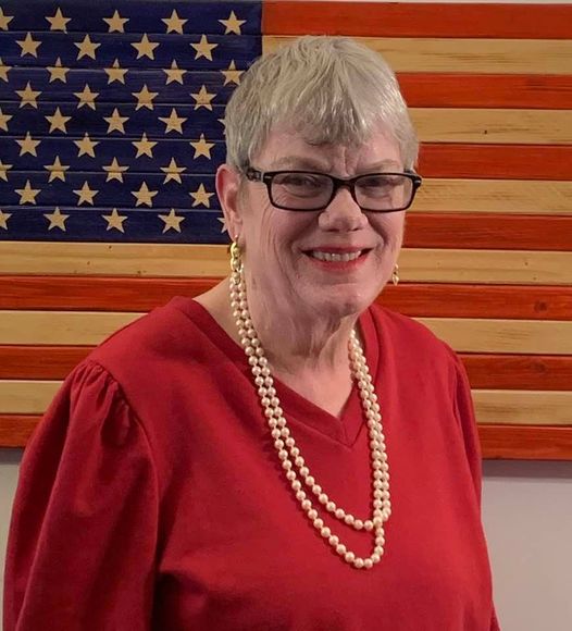 A woman in red shirt and pearls standing next to an american flag.